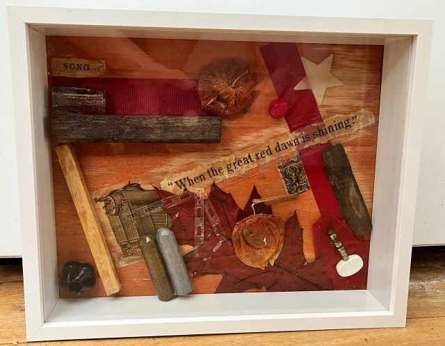 A framed shadow box artwork featuring various materials, including wood, metal, and fabric, arranged around a quote that reads, 'When the great red dawn is shining.'