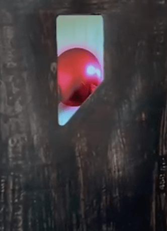 A close-up of a rectangular opening in a dark wooden surface, through which a shiny red ball is partially visible.